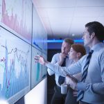 Three business people discuss graphs on screen in meeting room