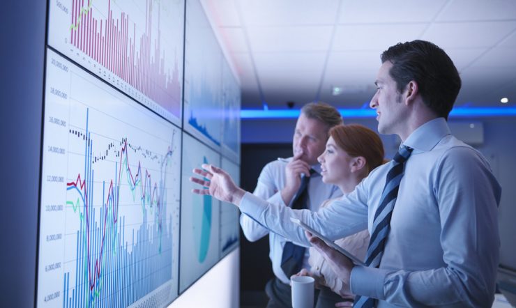 Three business people discuss graphs on screen in meeting room