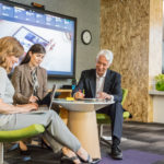 Group of two female and one male office workers brainstorming in informal office setting. Both women are using laptops while the man is writing. Large screen shown in background.