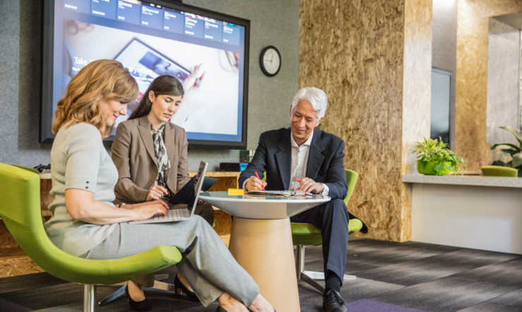 Group of two female and one male office workers brainstorming in informal office setting. Both women are using laptops while the man is writing. Large screen shown in background.