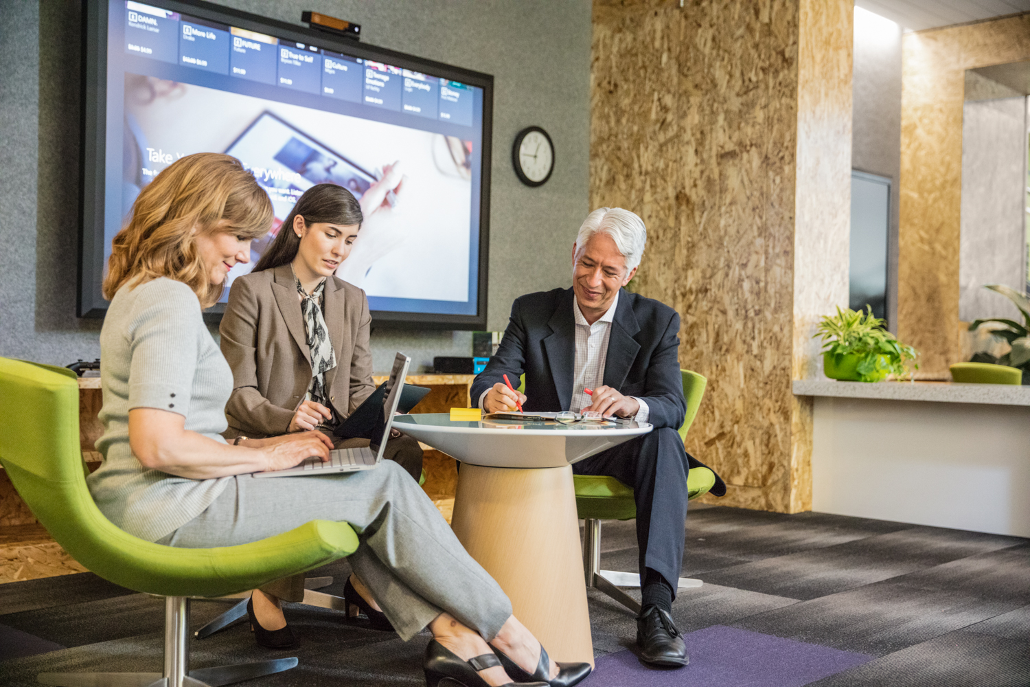 Group of two female and one male office workers brainstorming in informal office setting. Both women are using laptops while the man is writing. Large screen shown in background.