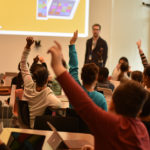 Een groep kinderen die een vinger omhoog steken om antwoord te geven aan de leraar.