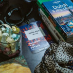 Uma coleção de livros de viagens, um badge, um copo com notas dentro, um lenço e uma caneca