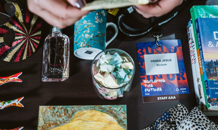 Umas mãos a agarrar um papel no ar e por baixo encontram-se uma máscara, um perfume, uma caneca, um copo comnotas dentro, um badge, uma coleção de livros de viagem, uma coleção de sardinhas em porcelana, um livro de pinturas e um lenço