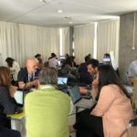 Vários grupos de pessoas sentados em mesas numa sala a participar no hackathon