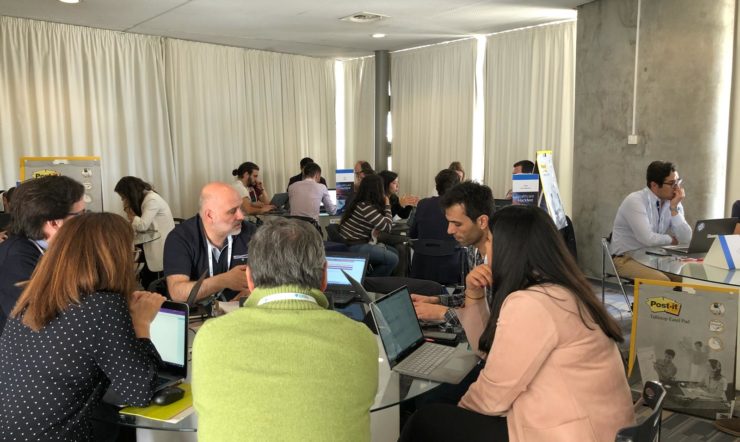 Vários grupos de pessoas sentados em mesas numa sala a participar no hackathon