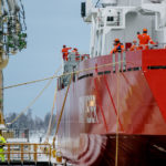 A large red ship with a few men onboard stationed at the shipyard