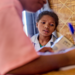 A female child sitting and looking as a nurse writes in notebook in rural clinic of nutrition enhancement program in Madagascar.