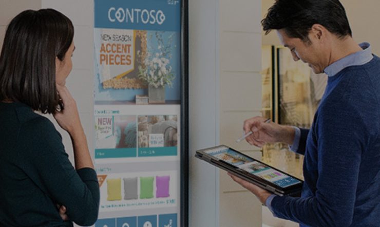 um homem a segurar um tablet e uma mulher de pé em frente a um ecrã digital a mostrar artigos da loja (almofadas)