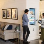 Female and male first line workers standing on sales floor in commercial retail store, in front of a wall-mounted monitor. She is using an Acer convertible laptop (folded open as a tablet) to navigate through images on monitor screen which shows product ads for home furnishings.