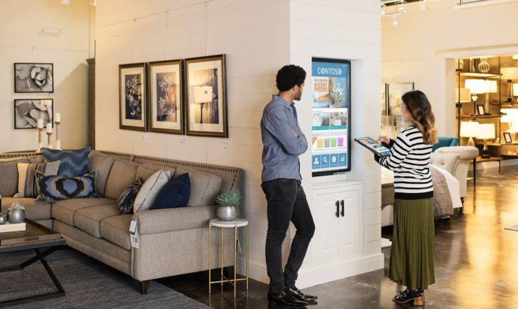 Female and male first line workers standing on sales floor in commercial retail store, in front of a wall-mounted monitor. She is using an Acer convertible laptop (folded open as a tablet) to navigate through images on monitor screen which shows product ads for home furnishings.
