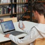 a man using a laptop computer sitting on top of a book shelf