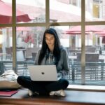 Female college student sitting cross-legged on windowsill using a Surface Book on her lap (screen not shown).