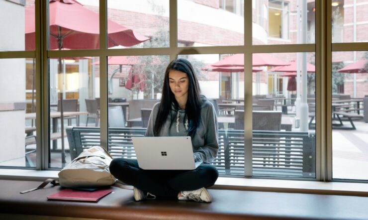 Female college student sitting cross-legged on windowsill using a Surface Book on her lap (screen not shown).