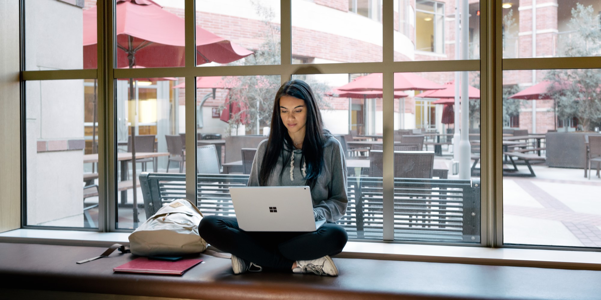 Female college student sitting cross-legged on windowsill using a Surface Book on her lap (screen not shown).