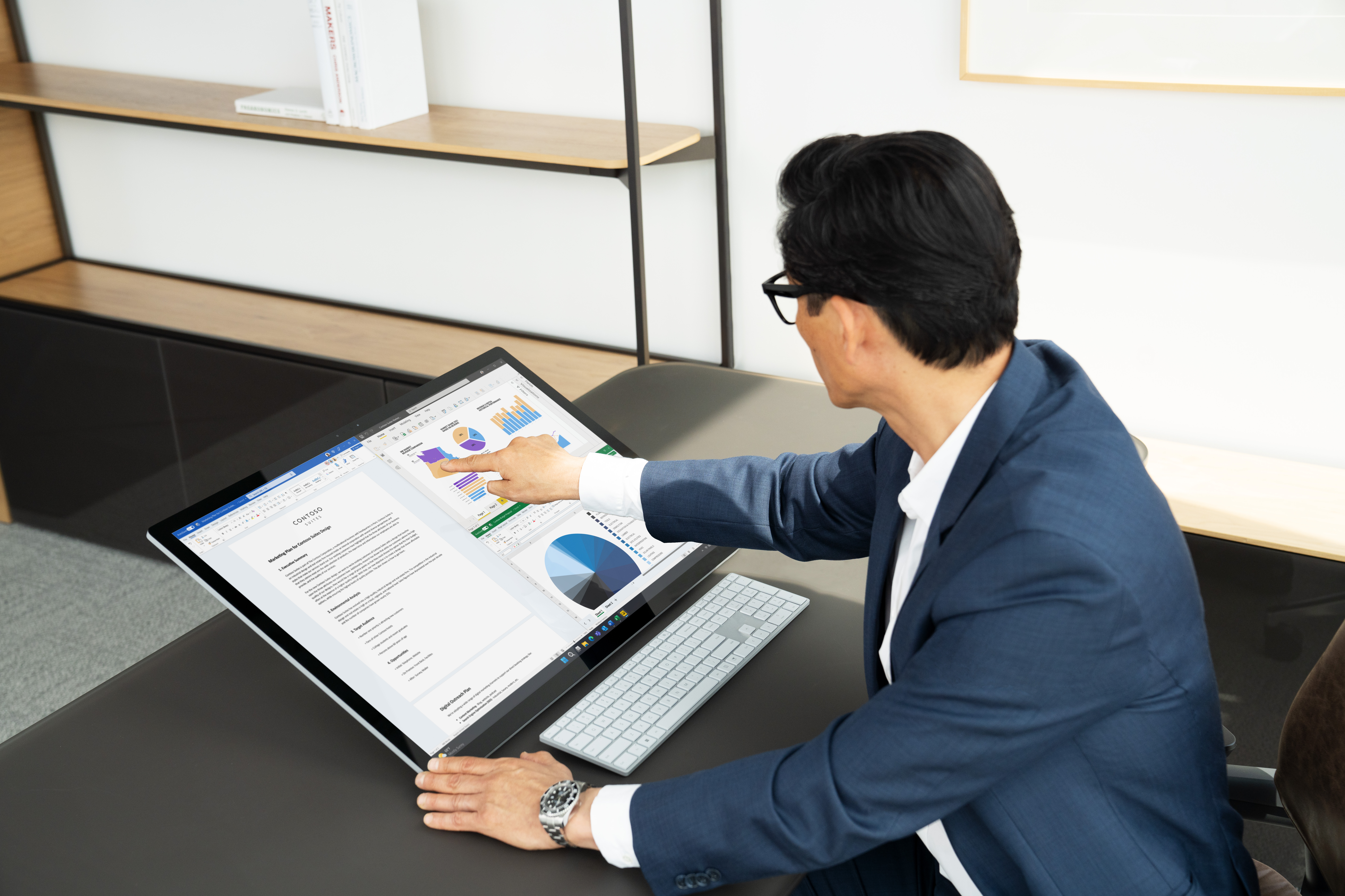 A man sitting at his office desk working on a Surface Studio 2 Plus showing Work, Excel and Power BI in Snap Assist.