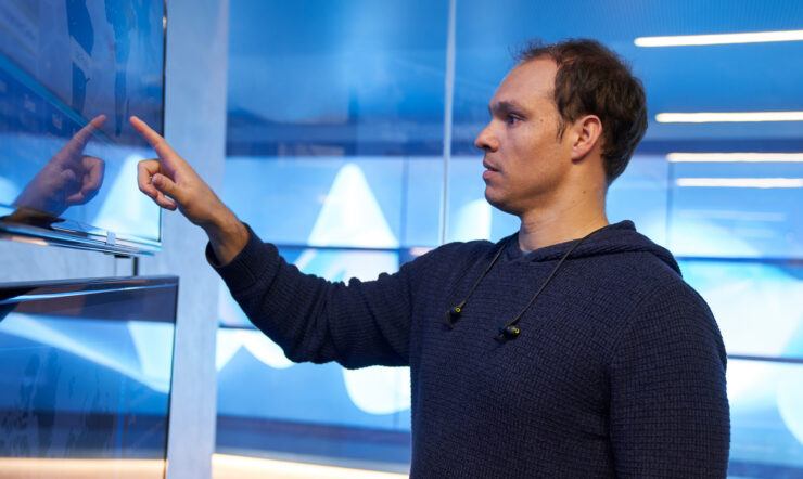 Man in a hooded sweater/sweatshirt inside a secure room, pointing at a geographic area displayed on a large monitor