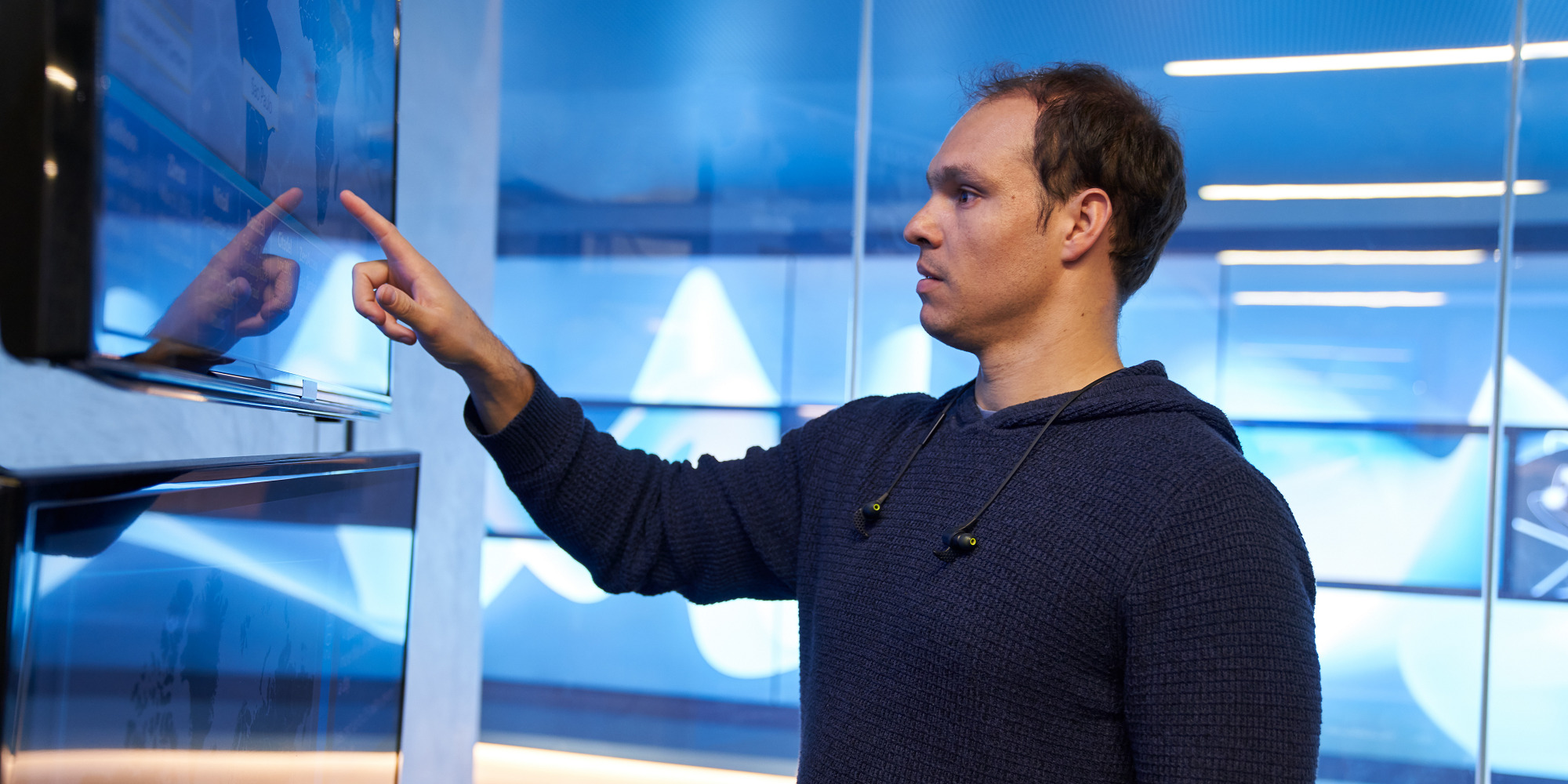 Man in a hooded sweater/sweatshirt inside a secure room, pointing at a geographic area displayed on a large monitor