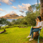 Woman in white t-shirt and jean on a swing working on a laptop with a lake an mountain in the background