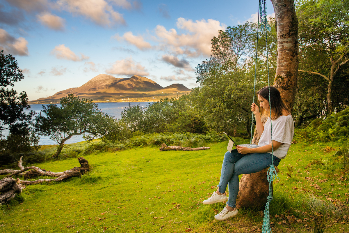 Woman in white t-shirt and jean on a swing working on a laptop with a lake an mountain in the background