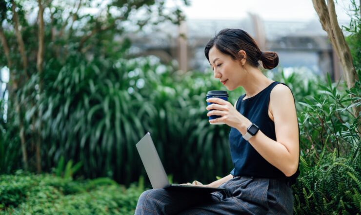 Young businesswoman uses her laptop outdoors, while sitting on a bench surrounded by green plants