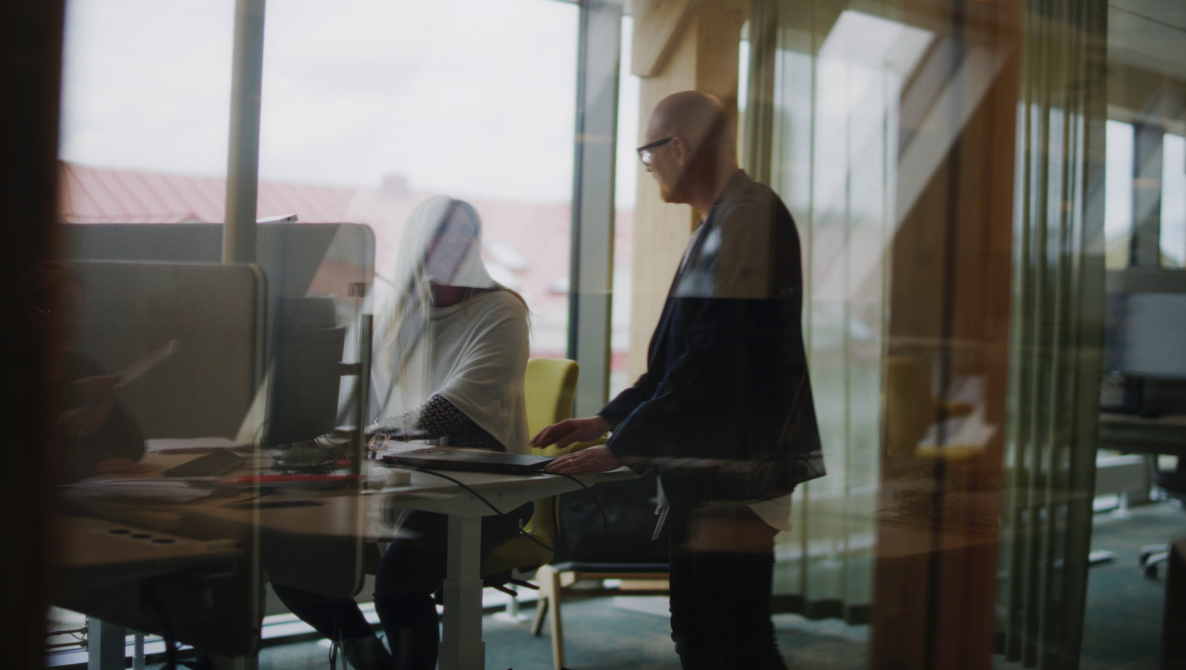 People in an office looking at screen, seen from behind a glass that is reflecting light.