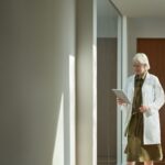 nainen valkoisessa lääkärin takissa nojaa neutraalin väriseen seinään pitäen tablettia kädessään.